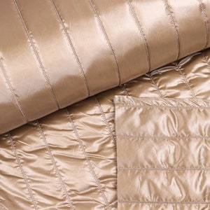 Può includere: Primo piano di un tessuto trapuntato beige con cuciture orizzontali. Il tessuto è liscio e lucido, con un leggero bagliore.