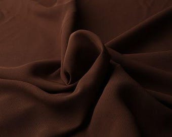 Op de afbeelding: Close-up van een vloeiende, donkerbruine stof met zachte plooien en subtiele textuur. De stof lijkt een lichtgewicht materiaal te zijn, mogelijk zijde of een vergelijkbaar drapebaar textiel. De belichting creëert schaduwen die de plooien benadrukken.