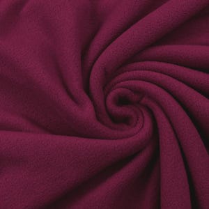 Könnte beinhalten: Nahaufnahme einer Wirbelung aus bordeauxfarbenem Fleece-Stoff. Der Stoff ist weich und strukturiert, mit leichtem Glanz. Die Falten erzeugen ein Spiralmuster, das die Tiefe und Textur des Materials hervorhebt. Ideal für Decken, Kleidung oder Bastelprojekte.
