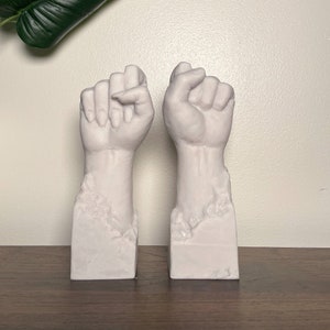 May include: Two white ceramic sculptures of raised fists, each with a textured base. The sculptures are on a wooden surface.