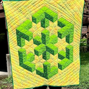 May include: A handmade quilt featuring a geometric design in shades of green and yellow. The quilt has a repeating pattern of cube-like shapes, with a green border. The background is a textured yellow fabric.