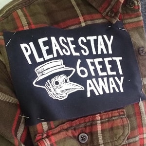 Może przedstawiać: Czarna łatka przyszyta do koszuli w kratę z napisem "PLEASE STAY 6 FEET AWAY" w białym kolorze. Pod tekstem znajduje się czarno-biała ilustracja lekarza od zarazy z długim dziobem.
