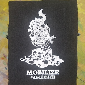 Può includere: Toppa in tessuto nero con una grafica bianca di una farfalla in fiamme su un blocco con il testo "MOBILIZE #AbolishICE"