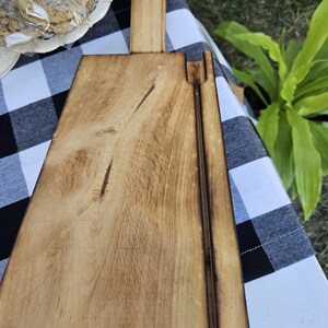 May include: A wooden cutting board with a hinged lid. The board is made of light wood and has a natural finish. The lid is closed and the board is sitting on a black and white checkered tablecloth.