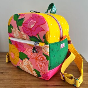 May include: A small backpack featuring a pink floral and butterfly print on the front pocket, a yellow polka-dot top panel, and a green side panel with a small white label reading Seu Happy. It includes yellow adjustable straps and silver zippers.