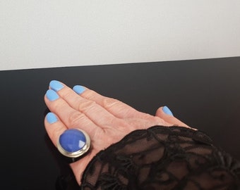 May include: A hand with light blue nail polish is wearing a silver ring with a large blue oval stone. The hand is resting on a black surface.