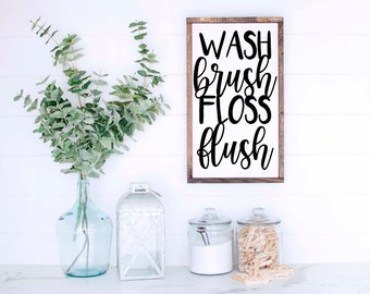 May include: A white wooden sign with black calligraphy that reads "WASH brush FLOSS flush". The sign is hanging on a white wall with white wood paneling. There is a vase with green eucalyptus branches, a white lantern, and two glass jars on a white countertop.