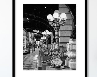 Peut inclure: Photographie en noir et blanc d'une rangée de lampadaires ornés de plusieurs lumières, bordant un trottoir sous une arche. Les lampadaires sont en métal et présentent un design décoratif. Le trottoir est mouillé et il y a des ombres des lampadaires.