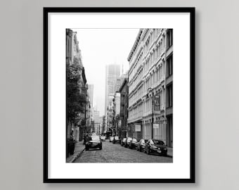 May include: Black and white photograph of a narrow street in a city with tall buildings on either side. The street is lined with parked cars and the buildings have many windows.