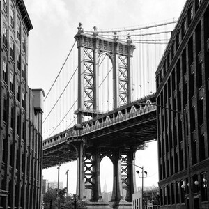 May include: A black and white photo of a bridge in New York City. The bridge is a suspension bridge with a large arch in the middle. The bridge is seen from a street below, with cars parked on both sides of the street. The bridge is a popular tourist destination in New York City.