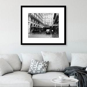 May include: A black and white photograph of a city street with a building in the background. A car is parked in the street and there are people walking by.
