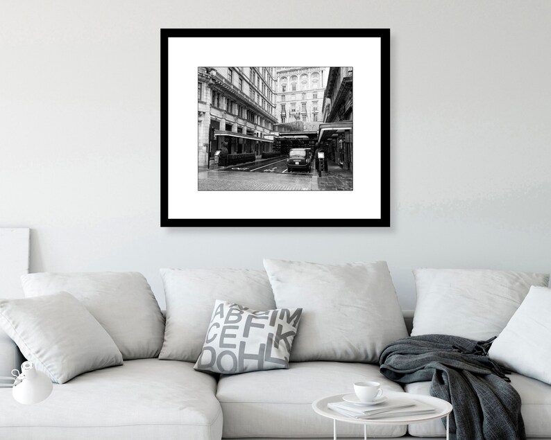 May include: A black and white photograph of a city street with a building in the background. A car is parked in the street and there are people walking by.