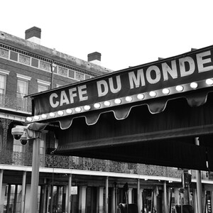 May include: Black and white photo of a cafe awning with the words "Cafe Du Monde" in white letters. The awning is lit with white lights and is in front of a brick building.