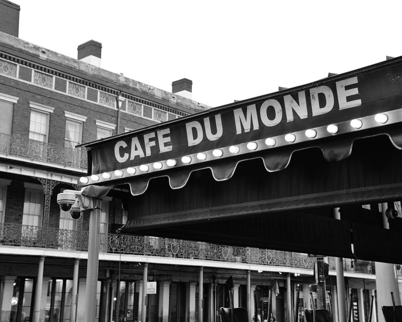 May include: Black and white photo of a cafe awning with the words "Cafe Du Monde" in white letters. The awning is lit with white lights and is in front of a brick building.