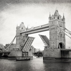 Op de afbeelding: Een zwart-witfoto van de Tower Bridge in Londen, Engeland. De brug is opgehaald, met een boot die eronderdoor vaart. De foto heeft een vintage, getextureerde uitstraling.