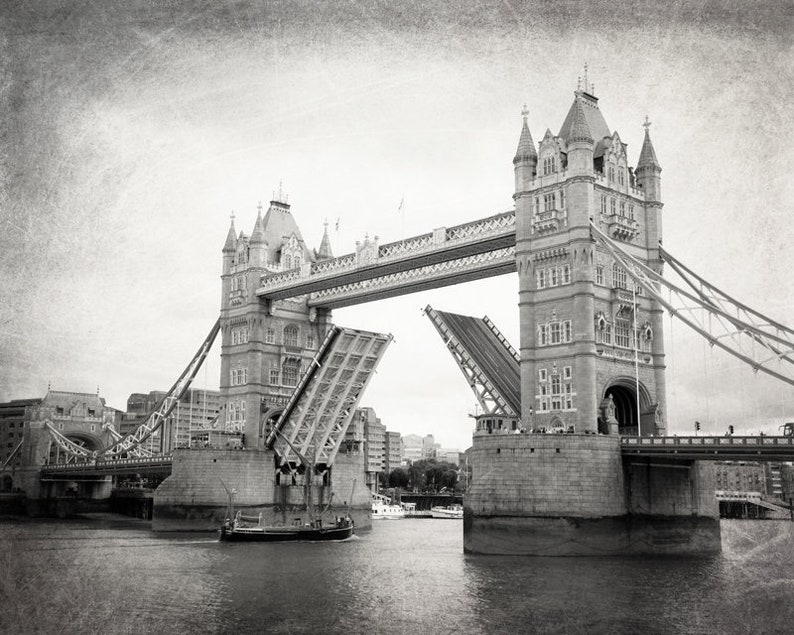 Op de afbeelding: Een zwart-witfoto van de Tower Bridge in Londen, Engeland. De brug is opgehaald, met een boot die eronderdoor vaart. De foto heeft een vintage, getextureerde uitstraling.