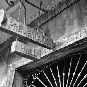 May include: Black and white photo of a sign that reads "Preservation Hall" with a smaller sign below that reads "Hall". The sign is hanging from a weathered wooden building with a wrought iron gate in the background.