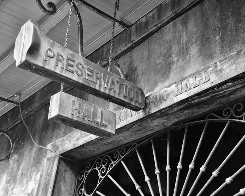 May include: Black and white photo of a sign that reads "Preservation Hall" with a smaller sign below that reads "Hall". The sign is hanging from a weathered wooden building with a wrought iron gate in the background.