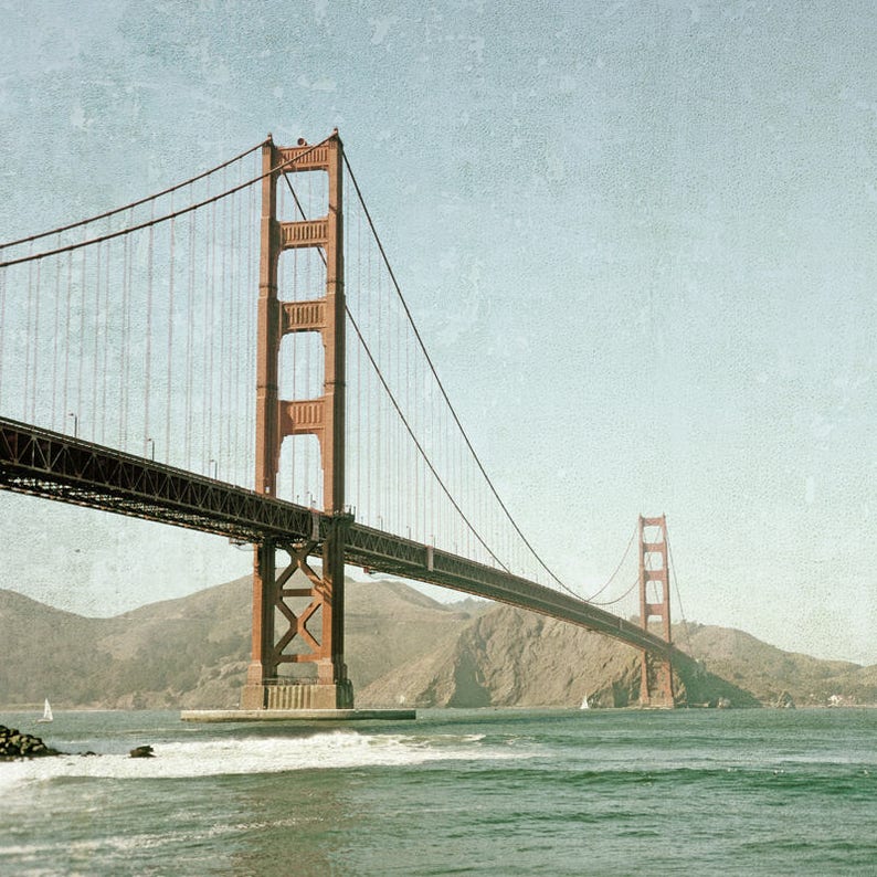 May include: A red suspension bridge with a large tower on the left side and a smaller tower on the right side. The bridge spans a body of water with hills in the background.