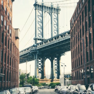 May include: A view of the Manhattan Bridge in New York City, seen from a street lined with brick buildings. The bridge is a steel structure with a large archway in the center. The street is lined with parked cars.