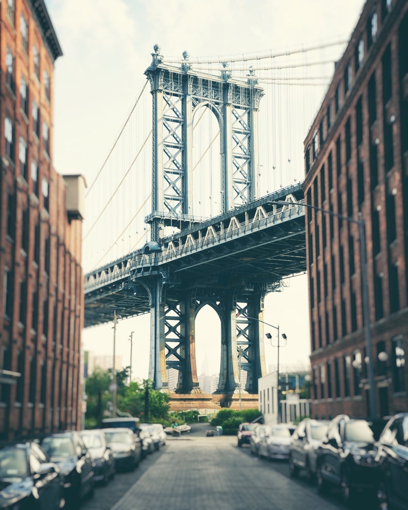 May include: A view of the Manhattan Bridge in New York City, seen from a street lined with brick buildings. The bridge is a steel structure with a large archway in the center. The street is lined with parked cars.