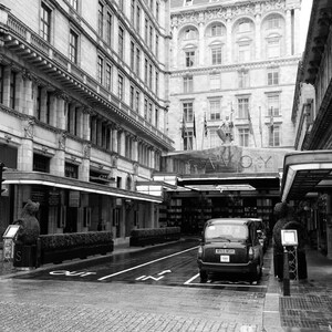 May include: A black and white photo of the Savoy Hotel in London, England. The hotel entrance is visible with a black taxi cab parked in front of it. The hotel sign reads "Savoy".