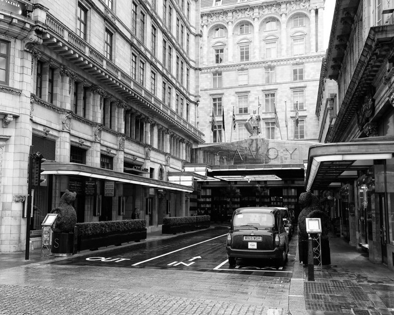 May include: A black and white photo of the Savoy Hotel in London, England. The hotel entrance is visible with a black taxi cab parked in front of it. The hotel sign reads "Savoy".