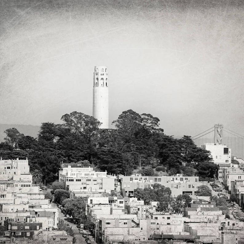 May include: A black and white photograph of a tall, white tower with a pointed roof, surrounded by trees and buildings. The tower is located in a city with a bridge in the background.