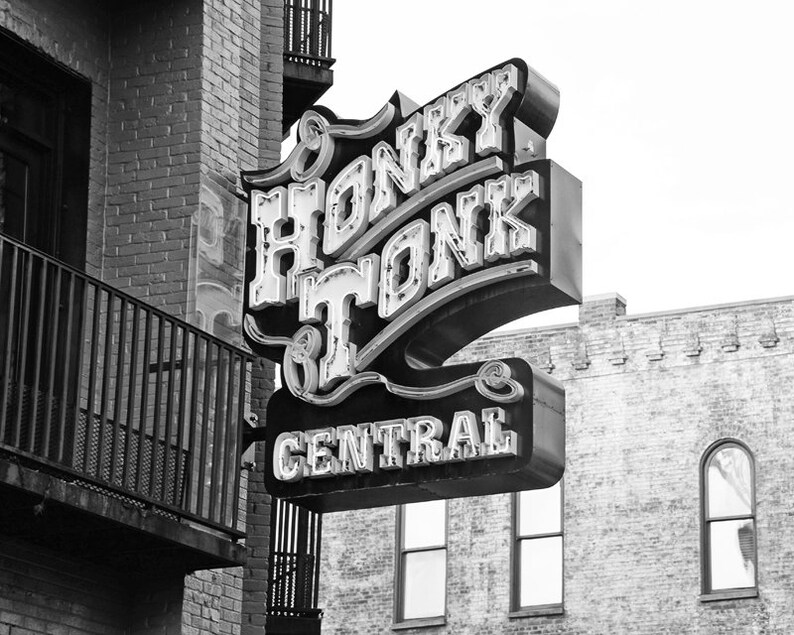May include: Black and white photograph of a neon sign that reads "Honky Tonk Central" in a retro font. The sign is mounted on a brick building.