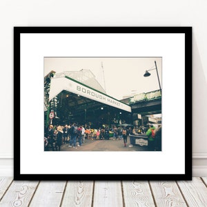 May include: Framed print of Borough Market, London. The image shows the market entrance with the words "BOROUGH MARKET" on a white awning. People are seen in the market, with a vintage aesthetic.