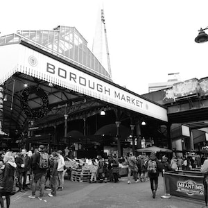 May include: A monochrome photograph of Borough Market, a busy food market. The market entrance is clearly visible, with the words "BOROUGH MARKET" displayed prominently. Numerous people are seen walking around the market. The Shard skyscraper is in the background.