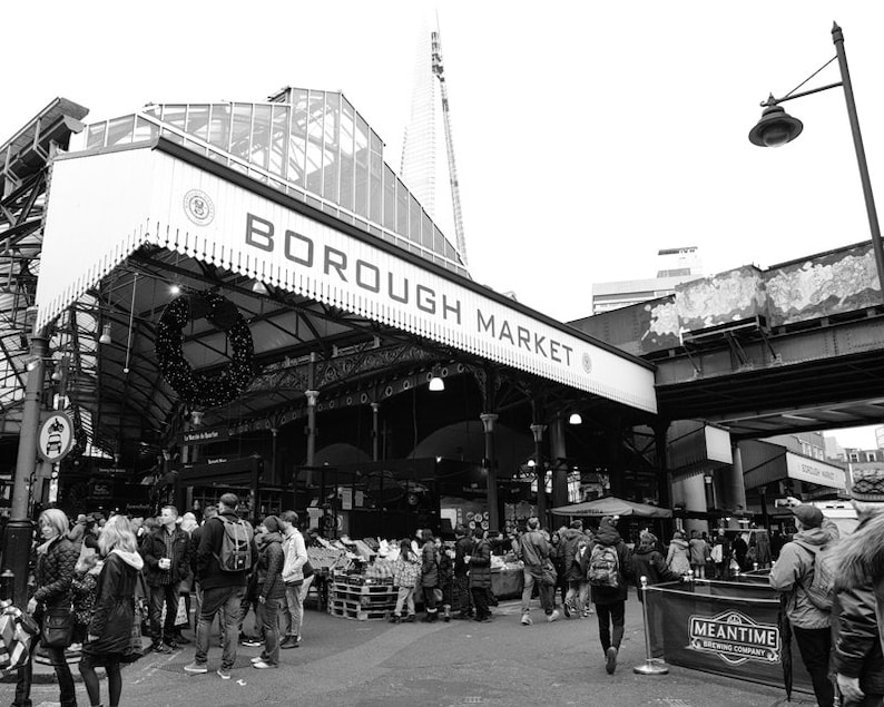 May include: A monochrome photograph of Borough Market, a busy food market. The market entrance is clearly visible, with the words "BOROUGH MARKET" displayed prominently. Numerous people are seen walking around the market. The Shard skyscraper is in the background.