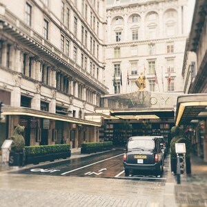 May include: A black London taxi cab is parked in front of the Savoy Hotel in London, England. The hotel is a historic landmark with a grand entrance and a sign that reads "Savoy".