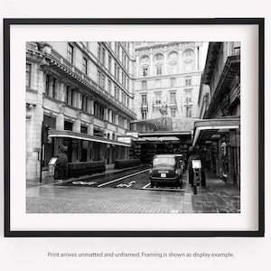 May include: A black and white photo of a London street with a black taxi cab parked in the middle of the street. The street is lined with buildings and there are people walking around.