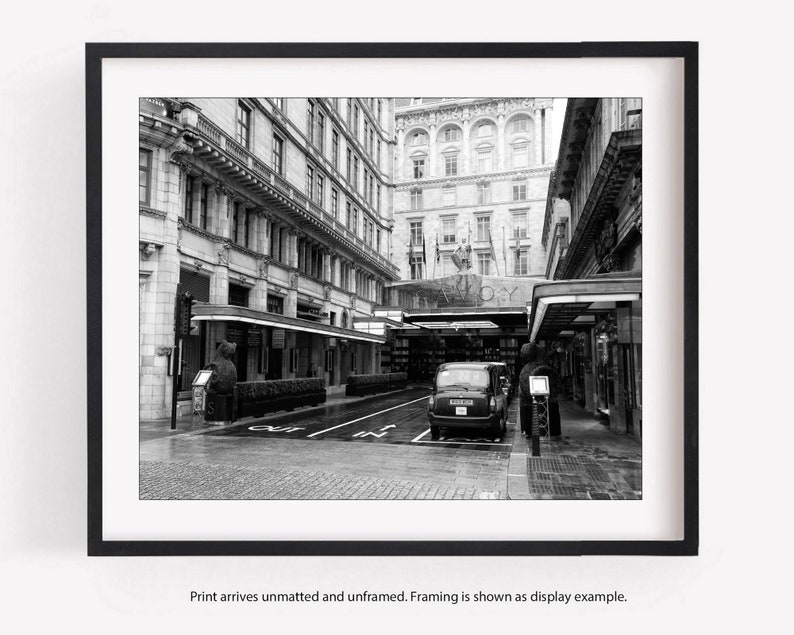 May include: A black and white photo of a London street with a black taxi cab parked in the middle of the street. The street is lined with buildings and there are people walking around.