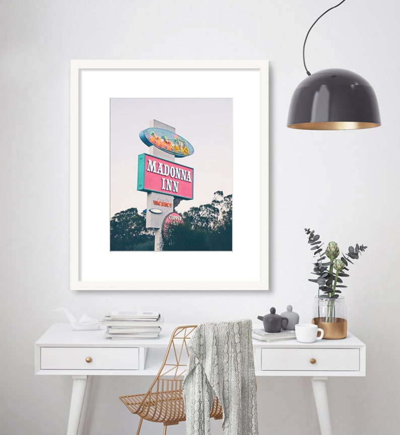 May include: A framed photograph of a vintage motel sign with the words "Madonna Inn" in pink and blue lettering. The sign is on a white pole with a red and white striped awning. The sign is set against a backdrop of trees and a cloudy sky.
