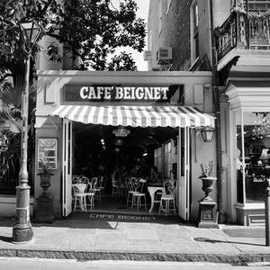 Op de afbeelding: Een zwart-witfoto van een caf&eacute; genaamd "Cafe Beignet" met een gestreepte luifel. Het caf&eacute; heeft tafels en stoelen buiten opgesteld. Op de voorgrond staat een lantaarnpaal en op de achtergrond een gebouw met een etalage.