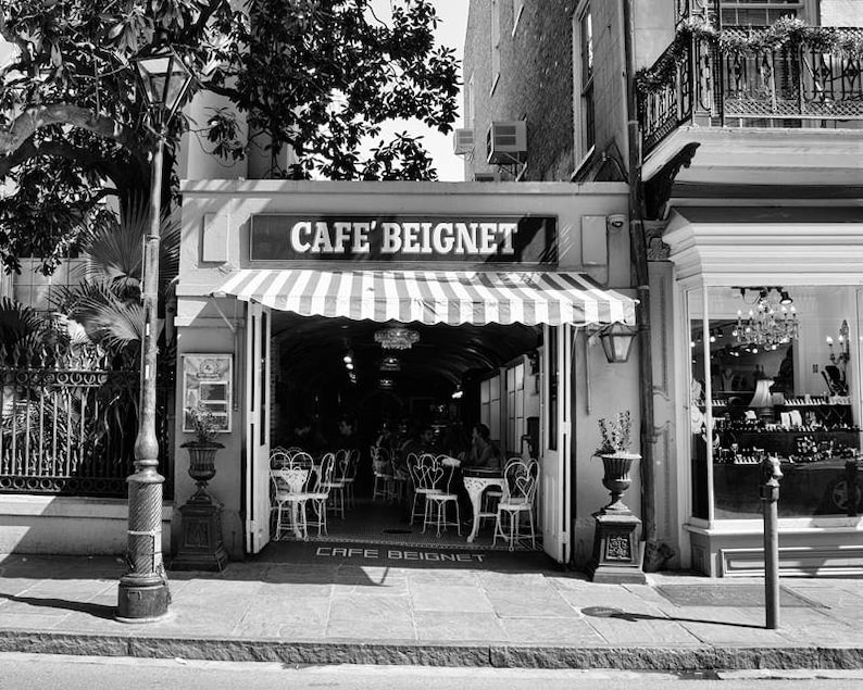 Op de afbeelding: Een zwart-witfoto van een caf&eacute; genaamd "Cafe Beignet" met een gestreepte luifel. Het caf&eacute; heeft tafels en stoelen buiten opgesteld. Op de voorgrond staat een lantaarnpaal en op de achtergrond een gebouw met een etalage.
