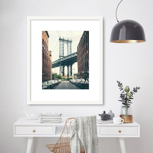 May include: A framed photograph of a bridge in a city. The bridge is made of metal and has a gray color. The bridge is surrounded by buildings. The photo is in a white frame.