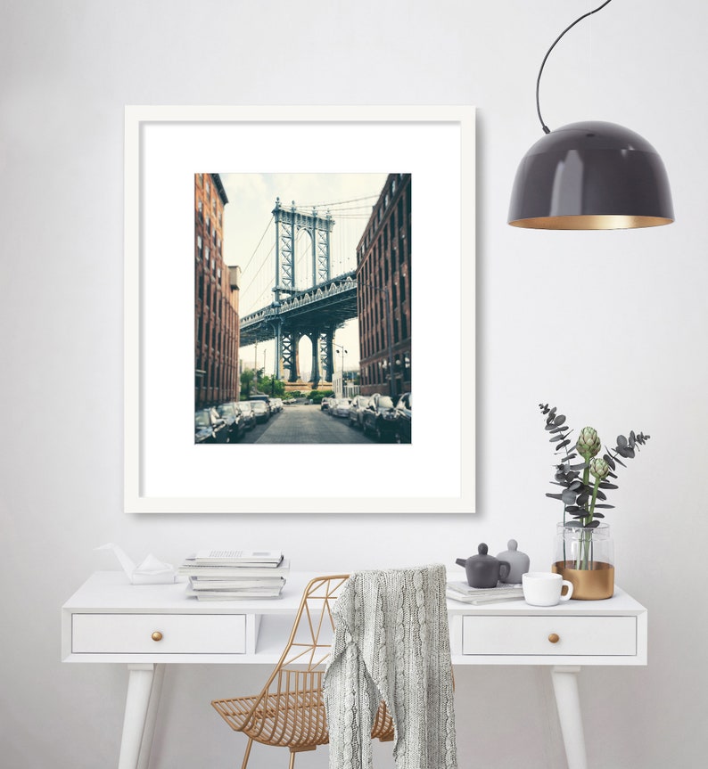 May include: A framed photograph of a bridge in a city. The bridge is made of metal and has a gray color. The bridge is surrounded by buildings. The photo is in a white frame.