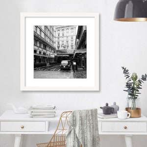 May include: Black and white photograph of a city street with a car parked in the middle of the road. The street is lined with buildings on both sides. The photo is framed in a white frame.