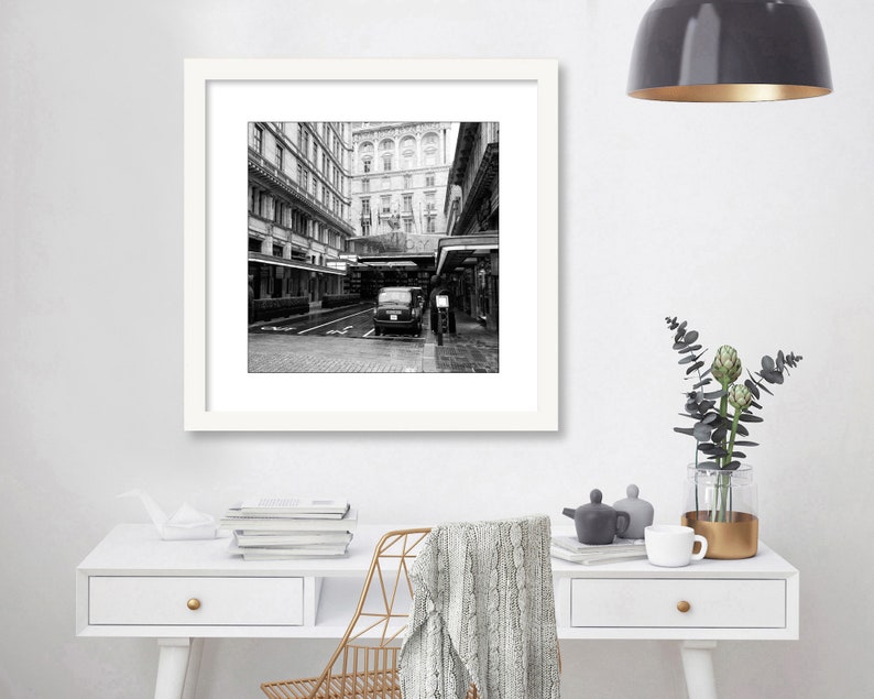 May include: Black and white photograph of a city street with a car parked in the middle of the road. The street is lined with buildings on both sides. The photo is framed in a white frame.