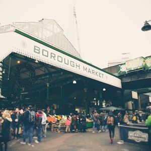 May include: A busy scene at Borough Market, London, with a white and green awning displaying the name in green letters. Shoppers browse stalls under the awning. A Christmas wreath hangs from the structure. The Shard is visible in the background.