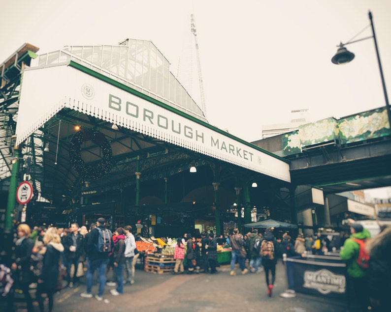 May include: A busy scene at Borough Market, London, with a white and green awning displaying the name in green letters. Shoppers browse stalls under the awning. A Christmas wreath hangs from the structure. The Shard is visible in the background.