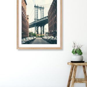 May include: A framed photograph of a street in New York City with a view of the Manhattan Bridge. The street is lined with brick buildings and parked cars. The bridge is a steel structure with a gray color.