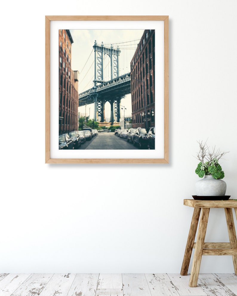 May include: A framed photograph of a street in New York City with a view of the Manhattan Bridge. The street is lined with brick buildings and parked cars. The bridge is a steel structure with a gray color.