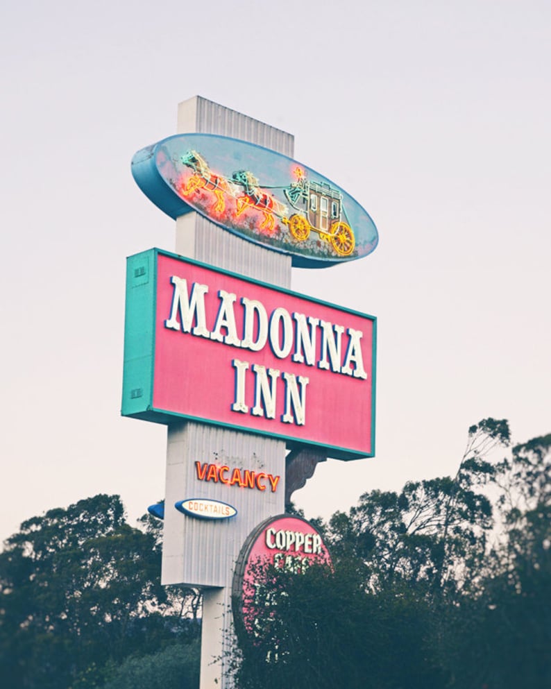 May include: A pink and blue neon sign with the text "Madonna Inn" in white letters. The sign is mounted on a white pole with a neon sign of a stagecoach with horses above the text. The sign is in front of a green tree.