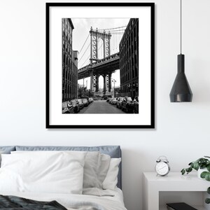 May include: A black and white photograph of a bridge in New York City. The bridge is seen from a street below, with cars parked on either side of the street.