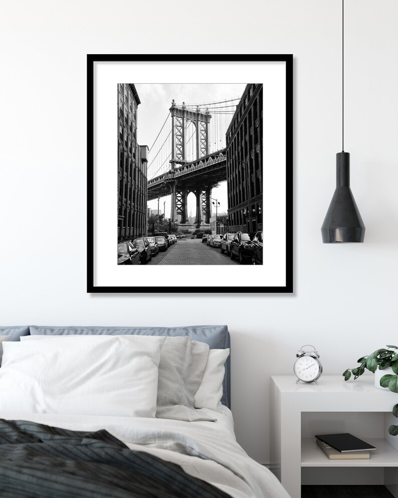 May include: A black and white photograph of a bridge in New York City. The bridge is seen from a street below, with cars parked on either side of the street.