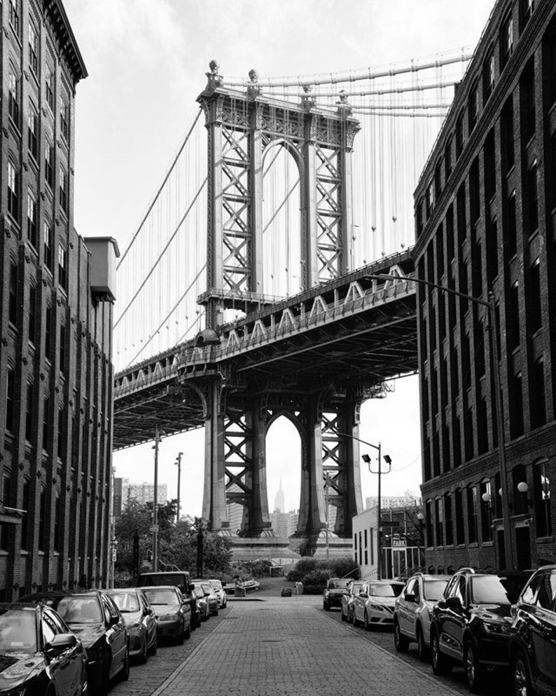 May include: A black and white photo of a bridge in New York City. The bridge is a suspension bridge with a large arch in the middle. The bridge is seen from a street below, with cars parked on both sides of the street. The bridge is a popular tourist destination in New York City.